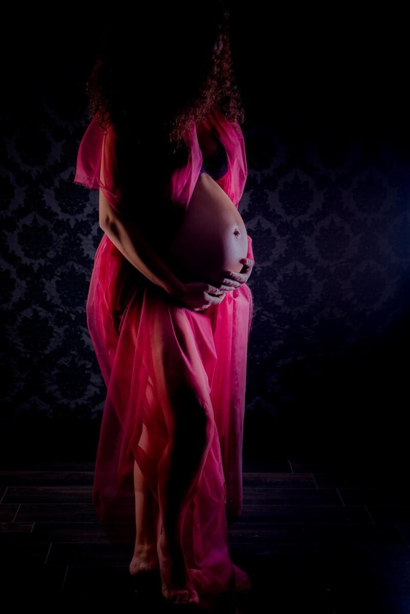 Une femme enceinte en robe rose posant devant un fond sombre au Studio gRossesse à Narbonne.
